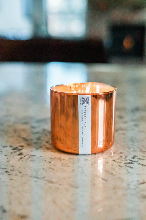 The Shanti + Tula Copper Vessel 3-Wick Soy Candle, featuring a white label and clean-burning ingredients, glows warmly on a marbled countertop—luxurious, USA-made decor perfect for inviting comfort and relaxation.
