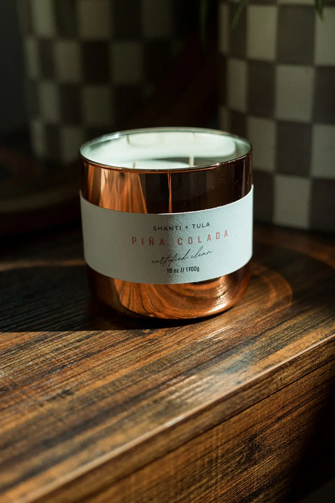 A Shanti + Tula Copper Vessel 3-Wick Soy Candle, labeled Pina Colada, sits on a wooden surface in sunlight. This USA-made, clean-burning luxury decor features exceptional fragrance with a checkered planter blurred in the background.