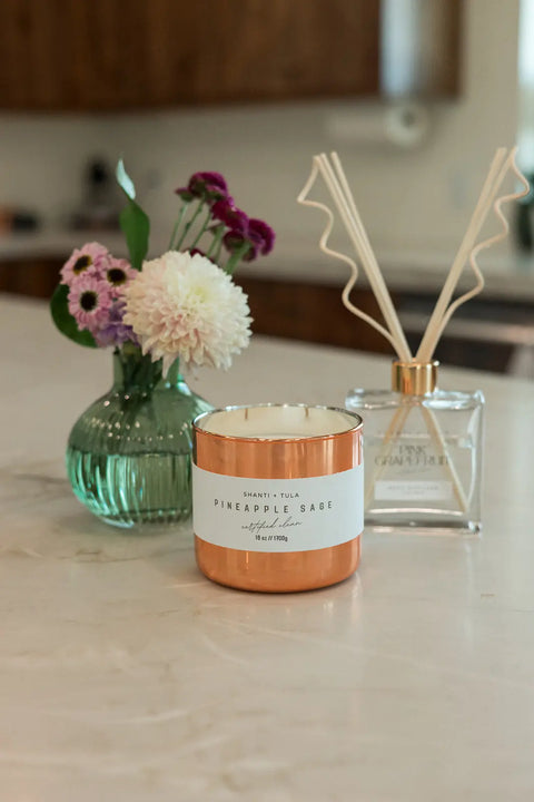 A Shanti + Tula Copper Vessel 3-Wick Soy Candle sits on a countertop near a green flower vase and reed diffuser, lending luxury decor and clean fragrance to the kitchen with this USA-made, gift-ready piece.