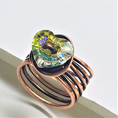 The Copper Wire Wrapped Crystal Heart Glam Ring by Alexa Martha Designs features a handmade copper wire wrap and a yellow iridescent Swarovski crystal heart centerpiece, shown on a light background.