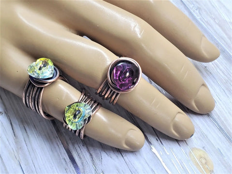 A mannequin hand displays three Copper Wire Wrapped Crystal Heart Glam Rings by Alexa Martha Designs—yellow-green rings on the pinky and middle fingers, and a purple one on the ring finger—against a light wooden background.