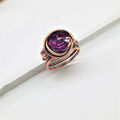 The Copper Wire Wrapped Crystal Heart Glam Ring by Alexa Martha Designs showcases a faceted purple gemstone set in an elegant copper wire wrap, beautifully displayed on a white surface with a diagonal line beneath it.