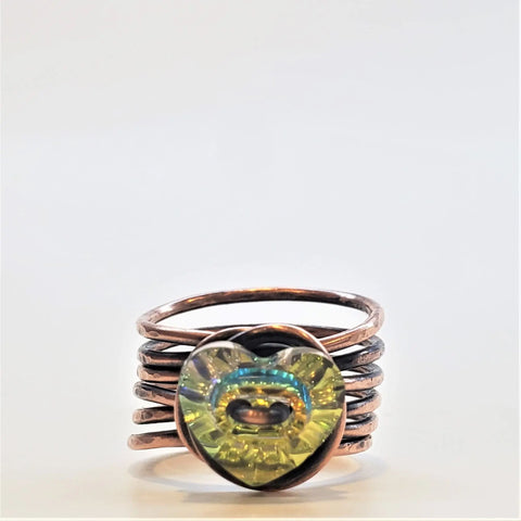 The Copper Wire Wrapped Crystal Heart Glam Ring by Alexa Martha Designs features stacked copper bands and an iridescent yellow-green glass heart centerpiece, capturing modern elegance against a plain white background.