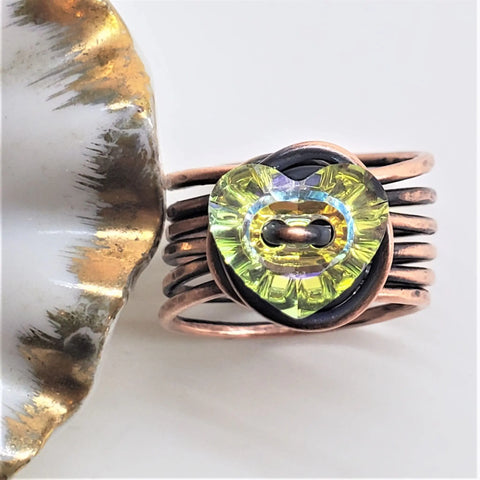The Copper Wire Wrapped Crystal Heart Glam Ring by Alexa Martha, from Alexa Martha Designs, features multiple copper bands and a yellow-green heart crystal centerpiece, displayed next to a gold-edged accent on a light background.