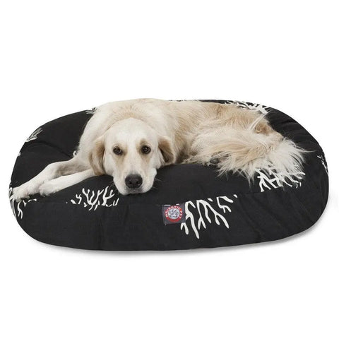 A light-colored dog lies on the Majestic Pet Coral Round Dog Bed, which is black with a white branch-like pattern and features a removable cover, looking forward with its head resting on the bed.