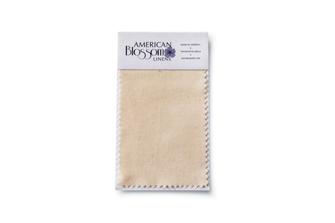 A rectangular beige fabric swatch made from USA Grown Cotton with zigzag edges is attached to a white paper label showing American Blossom Linens Swatches and product details, displayed on a white background.