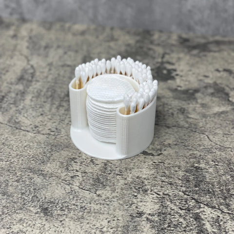 The Chatelet Manufacturing Cotton Swab Bathroom Organizer in a clean white finish keeps q-tips, cotton swabs, and exfoliating pads tidy and accessible, all displayed on a textured gray surface.
