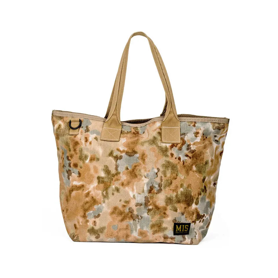 The MIS Tote Bag - Covert Desert features durable cordura nylon in a brown, green, and gray camo with tan handles. Water-resistant and made in the USA, it includes a black and yellow MIS logo patch on the bottom right corner.