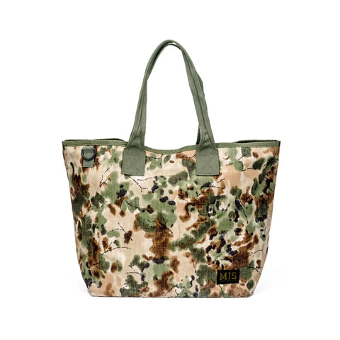 The MIS Tote Bag - Covert Woodland features durable Cordura Nylon in a green, brown, and beige camouflage pattern, sturdy green handles, a heavy-duty zipper closure, and a black and gold MIS patch near the bottom corner.