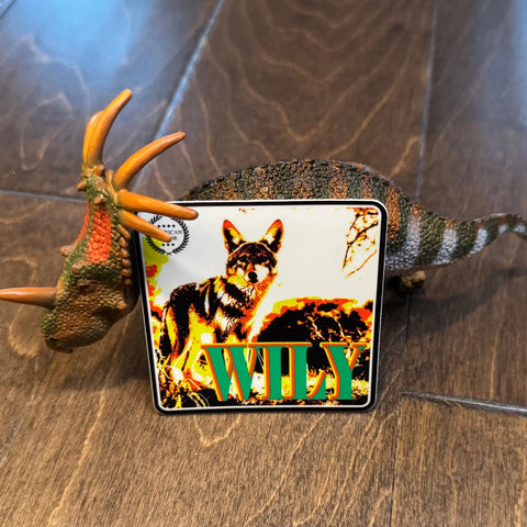 A toy triceratops stands on a wooden floor, holding a 3x3 square Sticker: Coyote - Wily from American Lictor, featuring a coyote image and the word WILY in bold green letters—a quirky accessory with forbidden doggo vibes.