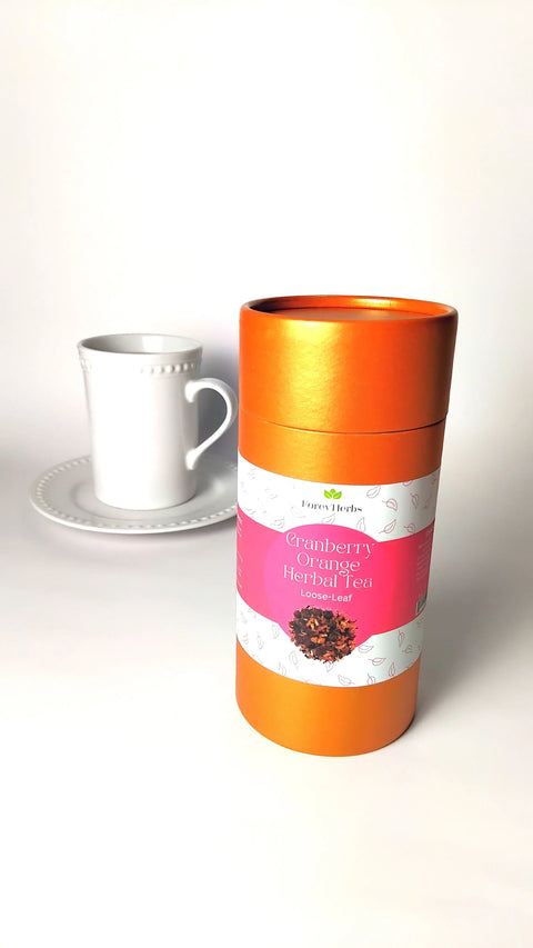 A cylindrical orange tin of Forev'Herbs Cranberry Orange Herbal Tea, a caffeine-free blend, is shown in front of a white teacup and saucer on a white background.