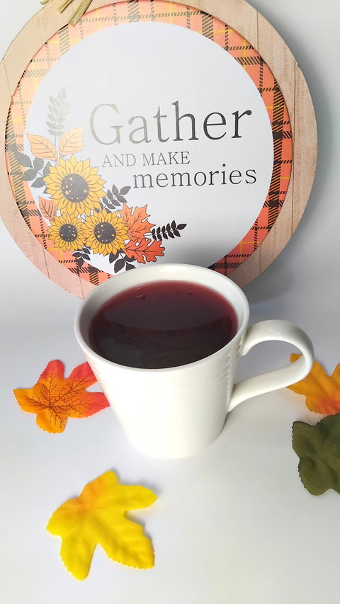 A white mug filled with Forev'Herbs Cranberry Orange Herbal Tea sits on a white surface, surrounded by artificial autumn leaves. Behind it is a decorative sign with sunflowers and plaid trim that reads, Gather and make memories.
