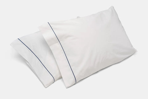 Two white Cotton Bed Pillowcases With Classic Piping Design by American Blossom Linens, featuring thin dark blue piping and made from 100% USA grown cotton, rest slightly overlapping on a white surface.