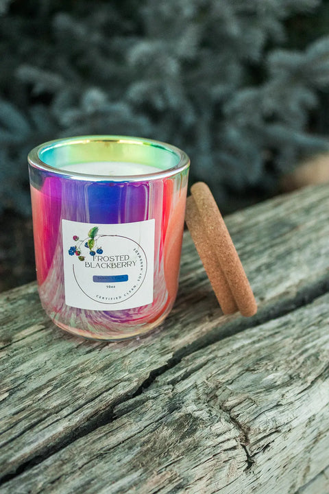 A 10 oz Cruel Summer Scented Soy Candle by Shanti + Tula, in a Prism glass jar with cork lid, sits on a rustic wooden surface. The USA-made candle features clean-burning soy wax, with greenery softly blurred in the background.