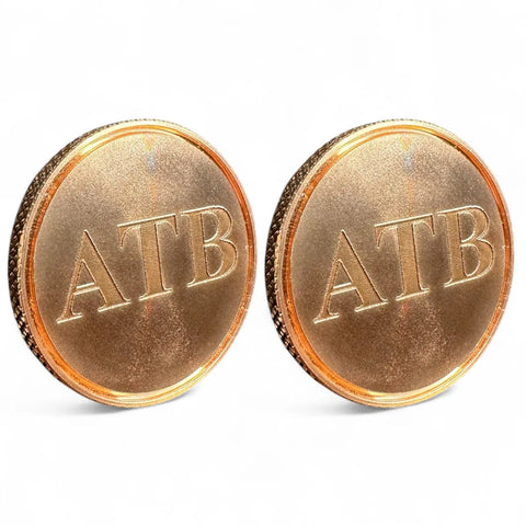 The Fairway Reserve Personalized Ball Markers (Set of 2) are upright with ATB engraved on each. Their shiny brass and copper tones stand out against a plain white background, resembling unique golf ball markers.