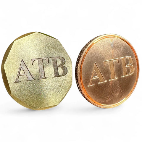Two coins, part of the Fairway Reserve Personalized Ball Markers (Set of 2), stand upright—a gold-toned brass marker with multiple edges and a round copper marker—each engraved with ATB, ideal for any golfer.