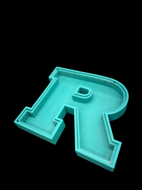 A turquoise, 3D letter R with a hollow, tray-like structure lies flat on a black background. This unique design by B. James Design & Woodwork is perfect for personalized Letter Piggy Banks.