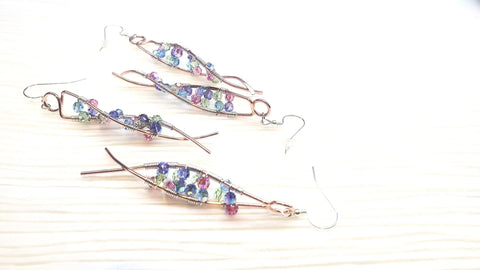 Four handmade wire earrings, including Alexa Martha Designs DNA Strand Double Helix Long Crystal Earrings with colorful beads, are displayed on a light wooden surface—perfect for science jewelry fans.