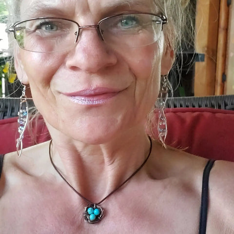 A woman with glasses and light blue eyes smiles gently, wearing Alexa Martha Designs DNA Strand Double Helix Long Crystal Earrings and a turquoise stone necklace, seated on a red cushion outdoors.
