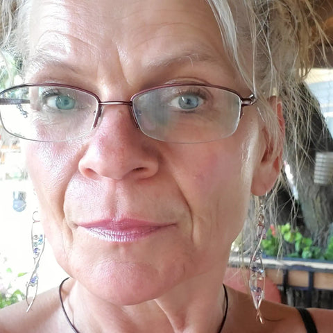 Close-up of an older woman with fair skin, glasses, light eyes, and lipstick, wearing Alexa Martha Designs DNA Strand Double Helix Long Crystal Earrings. Her hair is pulled back; the blurred background shows some outdoor elements.