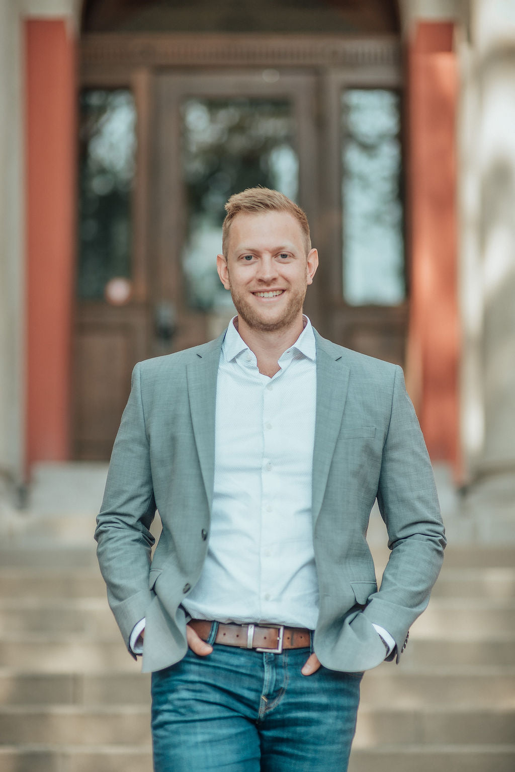 Man in a light gray blazer and blue jeans standing outdoors with a blurred background