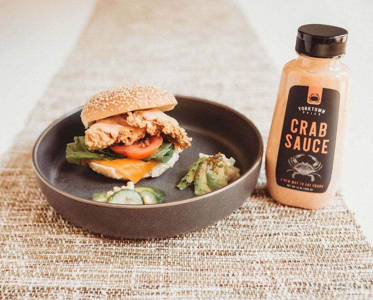 Burger with crab sauce on a plate next to a bottle of crab sauce on a woven mat.