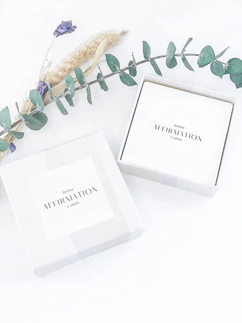 An open white Daily Affirmation Card Deck by Shanti + Tula rests on a light surface, its lid nearby. A green eucalyptus branch and dried flowers above the box create a mindful setting for inspiring, self-care messages. Made in the USA.