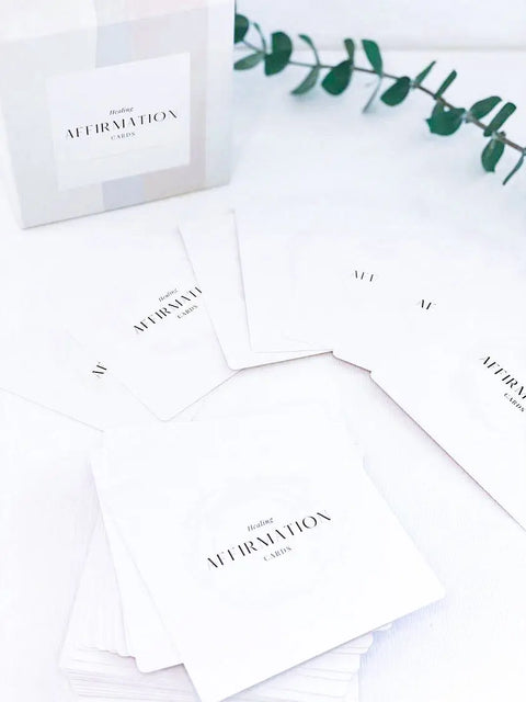 The Shanti + Tula Daily Affirmation Card Deck, a mindfulness and self-care gift made in the USA, is displayed with its box on a white surface, while a green leafy plant brings calm to the background.