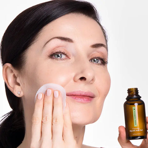 A woman with fair skin uses a cotton pad to apply Open Formula Dark Spot Remover Superset to her cheek. She has dark hair pulled back and is smiling slightly, treating hyperpigmentation against a white background.