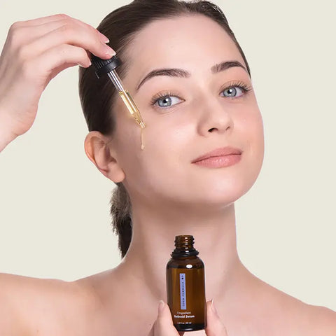 A woman applies Open Formula’s Dark Spot Remover Superset to her cheek with a dropper, holding the brown glass bottle, targeting hyperpigmentation and dark spots against a light background.