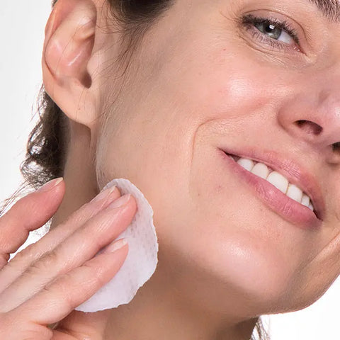 A woman smiles as she gently wipes her cheek with a cotton pad, applying Open Formula’s Dark Spot Remover Superset to target dark spots and hyperpigmentation. The close-up highlights her smooth, clean skin and focused skincare routine.