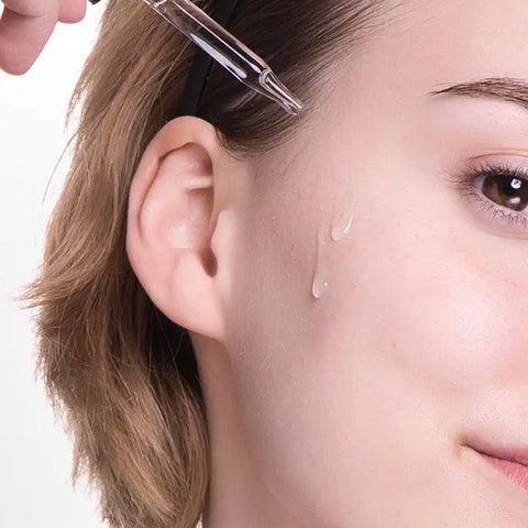 A person applies Open Formulas Dark Spot Remover Superset to their cheek with a dropper, targeting dark spots and hyperpigmentation. Only part of their face and ear are visible, with visible drops of the clear serum on their skin.