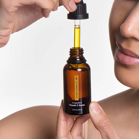 A person holds a brown glass dropper bottle of Open Formula Dark Spot Remover Superset and dispenses yellow serum near their face to target dark spots. The background is white, with the persons face partially visible.