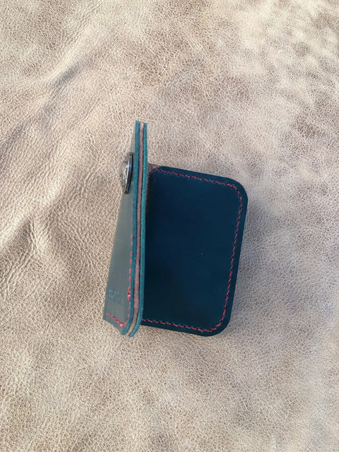The Sully + Proper Supply Deep Sea Teal Card Wallet, featuring red stitching, stands upright and slightly open on a textured tan leather surface.