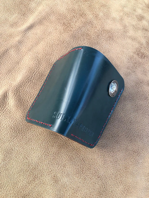 A Deep Sea Teal Card Wallet by Sully + Proper Supply with red stitching and a metal button rests on tan textured leather, featuring the brand’s name embossed on the front.