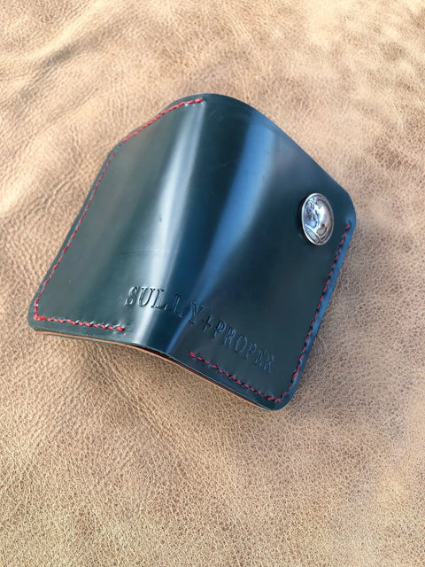 A Deep Sea Teal Card Wallet by Sully + Proper Supply, featuring handmade leather, red stitching, and a silver snap button, rests on a tan leather surface. The wallet is embossed with the brands name.