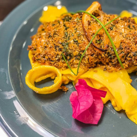 Part of the Deluxe Land & Sea Grilling Collection | Set of 4 by vendor-unknown, this dish showcases a breaded, herb-crusted fillet with all-natural spices over yellow pasta, topped with fried onion rings, chives, and an edible flower on a gray plate.