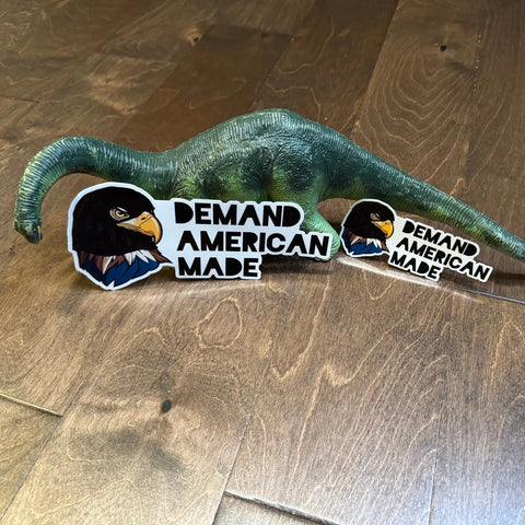 A green toy dinosaur sits on a wooden floor with two American Lictor Sticker: Demand American Made—perfect for adding to party favor bags or medium gift bags at your next celebration.