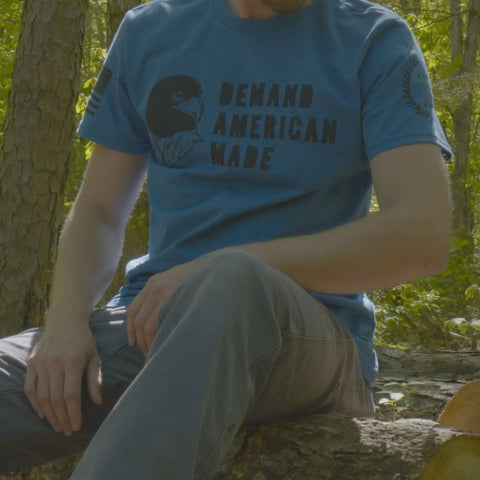 Outdoors on a tree trunk, a person wears the American Lictor T-Shirt: Demand American Made in blue with an eagle graphic and DEMAND AMERICAN MADE text. Face not visible. See our Size Guide to find your perfect fit.