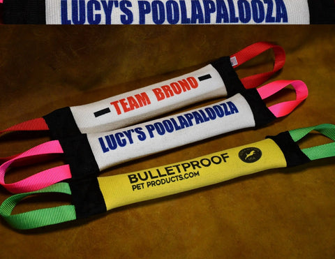 Three vibrant Dock Diving Bumper Tug - Competition Series Weighted toys by Bulletproof Pet Products Inc, each customizable with text like TEAM BRONO and LUCYS POOLAPALOOZA, are displayed on a brown surface with easy-grip handles.