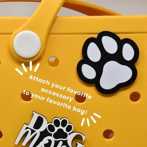 A yellow Bogg Bag features a Chatelet Manufacturing FRESHe BAGLETS Dog Paw Charm accessory with “DOG mom” text. “Attach your favorite tote bag accessory to your favorite bag!” appears in white across the bag.