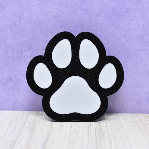The Chatelet Manufacturing FRESHe BAGLETS Dog Paw Charm, made in the USA, decorates Bogg Bags and other totes. Shown on light wood with a purple backdrop, this black and white charm is a cute accessory for your favorite tote bags.