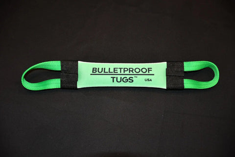 A green and black Doing Bad Dog Shit Fire Hose Training Tug (Bad Tug Line) by Bulletproof Pet Products Inc lies flat on a black surface, featuring two looped handles and a padded, branded center section.