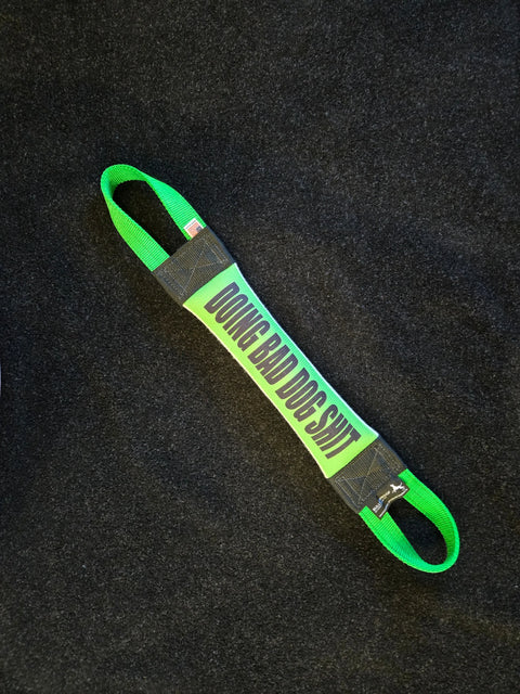 The Doing Bad Dog Shit Fire Hose Training Tug by Bulletproof Pet Products Inc is a green and black heavy-duty dog toy with “DOING BAD DOG SHIT” text, shown on a dark textured surface.