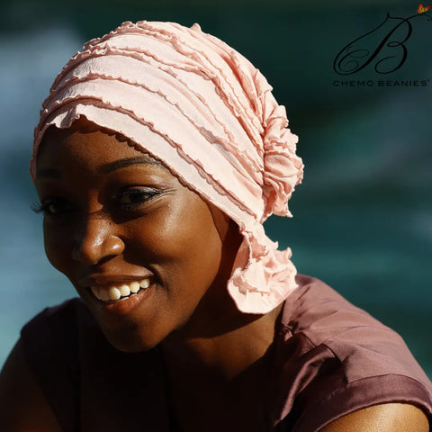 A woman smiles warmly in the sunlight, wearing a light pink Donna chemo beanie from Chemo Beanies® with a textured design, paired with a mauve top. The softly blurred background features a pool or water surface.