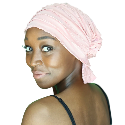 A woman with glowing skin and subtle makeup smiles softly over her shoulder, wearing the Chemo Beanies® Donna ruched pink chemo scarf and a black spaghetti strap top against a plain background.