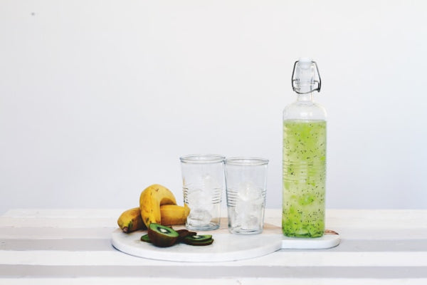 Bottle of green juice, two glasses, and a banana on a white surface with a white background