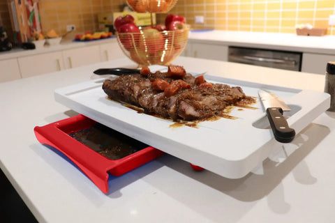A cooked steak with sauce and tomatoes sits on the Karving King Dripless Cutting Board 2 in 1 System, featuring a knife and red drip tray to collect juices. The scene is set in a modern kitchen with fruit and yellow tiles in the background.