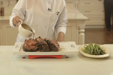 A chef in a white coat uses the Karving King Dripless Cutting Board 2 in 1 System to carve a roast, pouring sauce over the slices while asparagus rests on a plate nearby on the kitchen counter.
