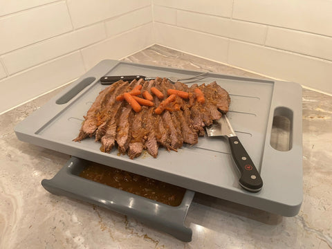 Sliced beef brisket and baby carrots sit on the Karving King Dripless Cutting Board 2 in 1 System, which features a built-in drip tray. Two knives rest on the board, placed on a marble countertop with a tiled backsplash.
